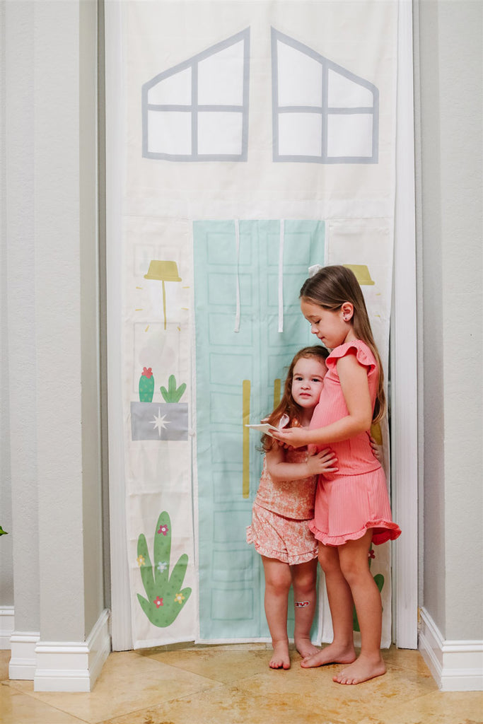 Two little girls hugging while playing with a doorway playhouse with a roll up blue door and decorated with cactuses. Sophisticated playroom design ideas